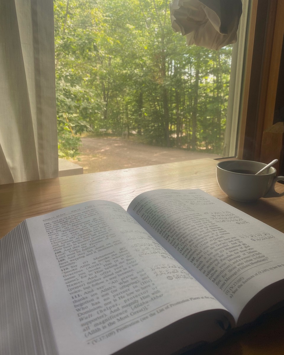 An open mushaf of the Qur'an by a sunlit window, its pages resting in soft light.